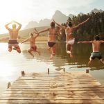 Portrait of young friends jumping from jetty into lake. Friends in mid air on a sunny day at the lake.